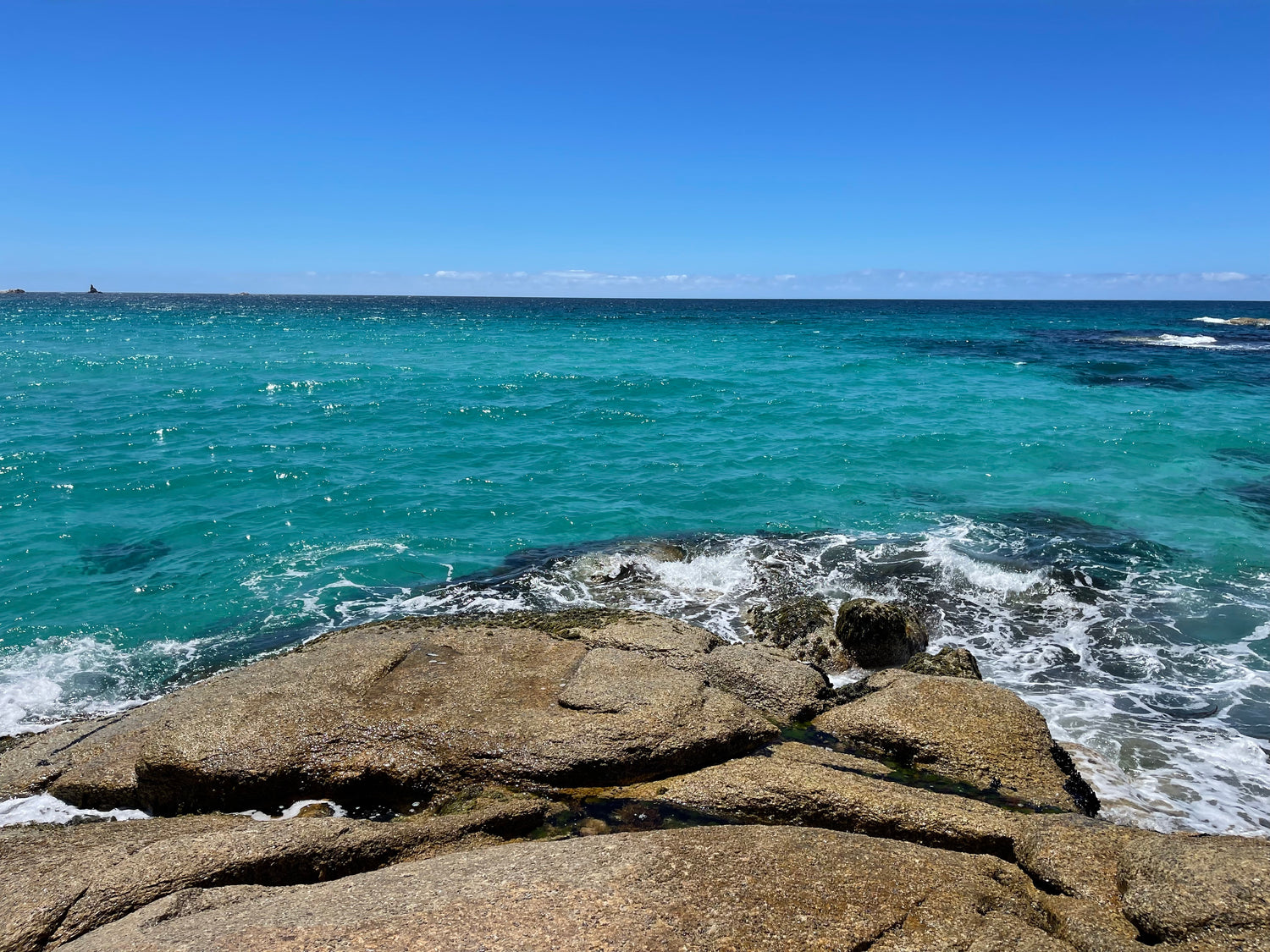 A photograph taken from some rocks on the beach foreshore. In the foreground are some brown rocks, and then crystal clear turquoise water, with a cloudless blue sky.